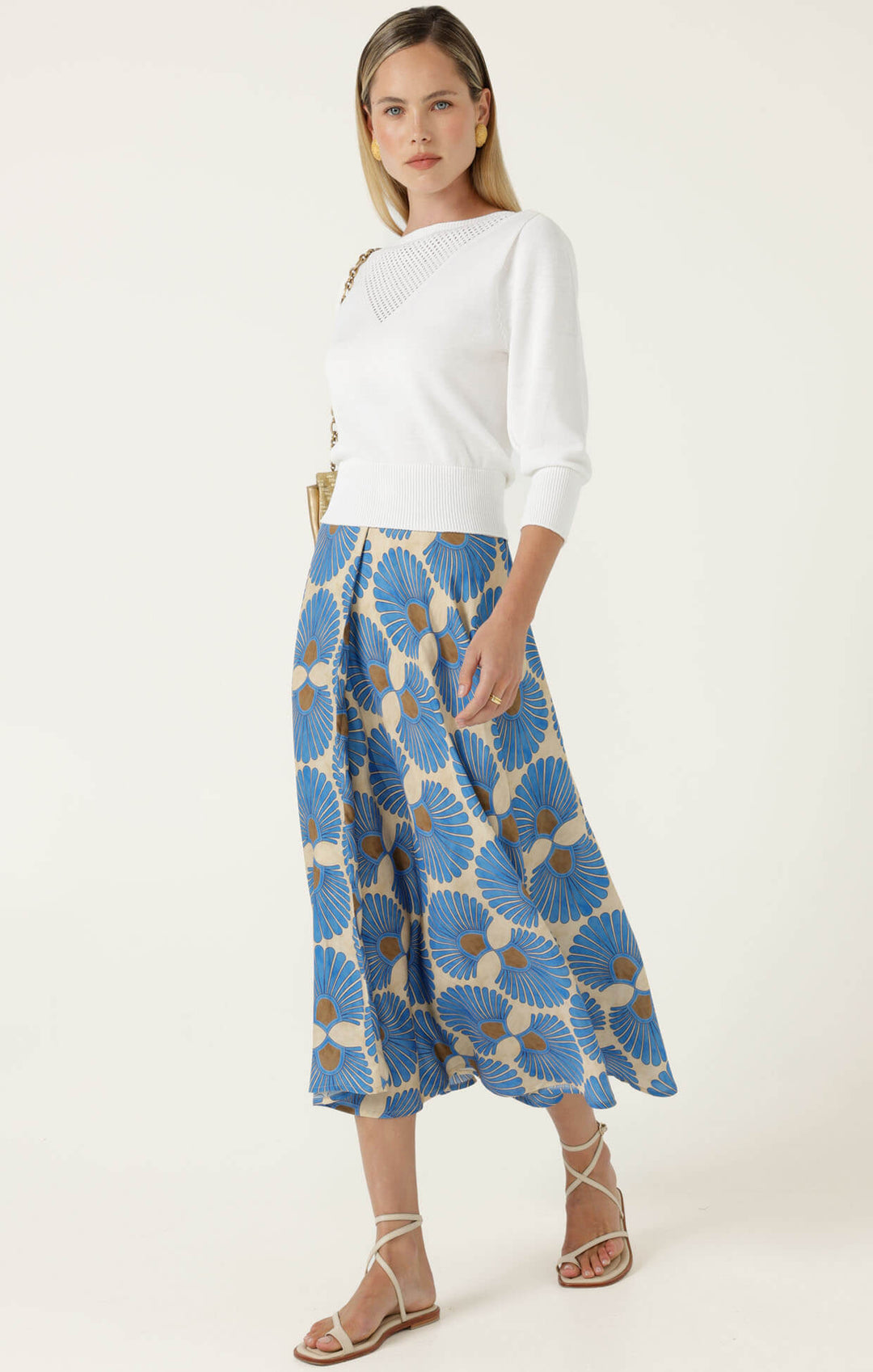 Sacha Drake  modeling a white sleeveless top with a textured chest panel and a high-waisted midi skirt featuring a blue, beige, and brown floral fan pattern, paired with beige strappy heels and holding a small metallic clutch. On a white studio background