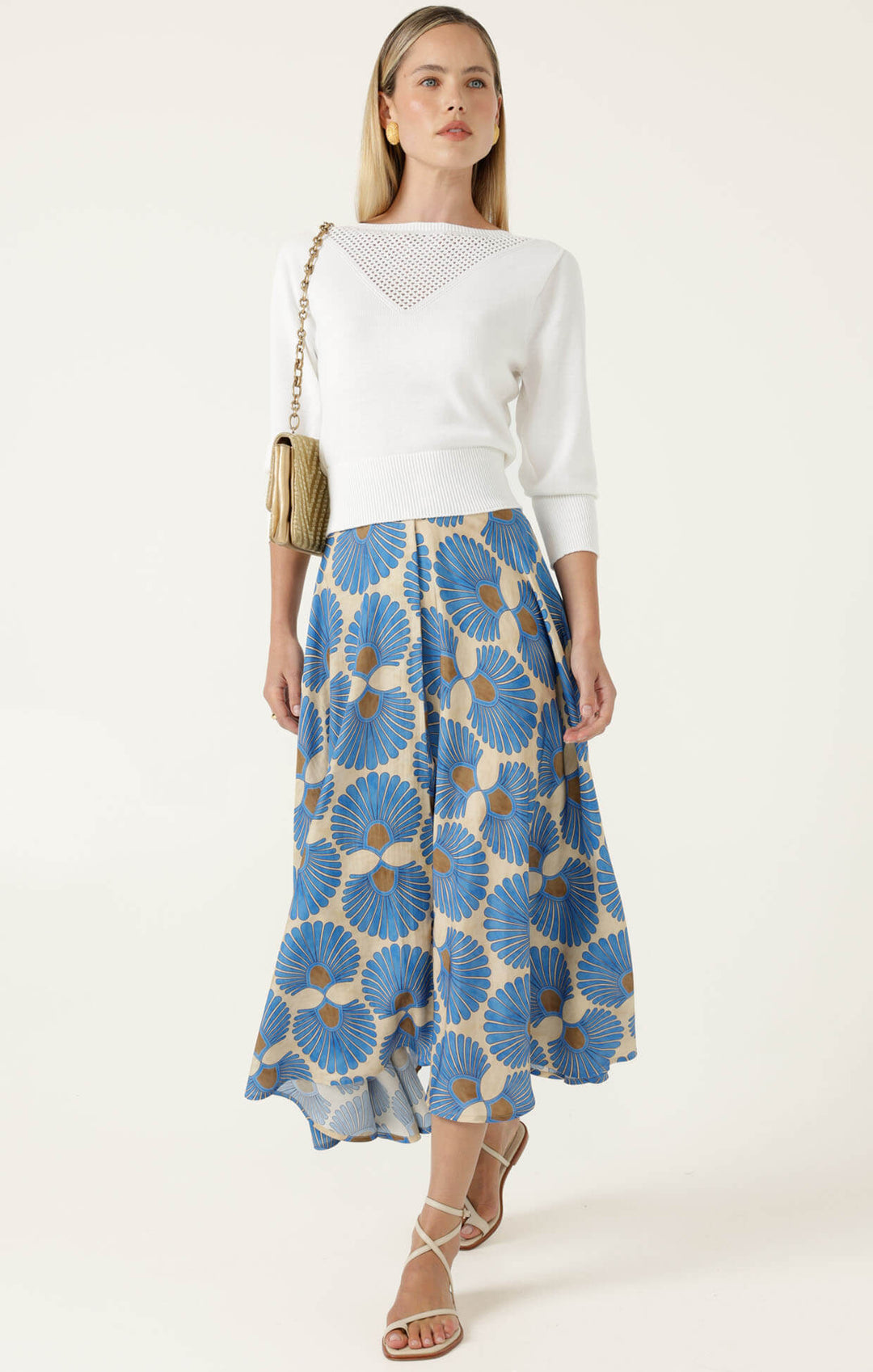 Sacha Drake  modeling a white sleeveless top with a textured chest panel and a high-waisted midi skirt featuring a blue, beige, and brown floral fan pattern, paired with beige strappy heels and holding a small metallic clutch. On a white studio background