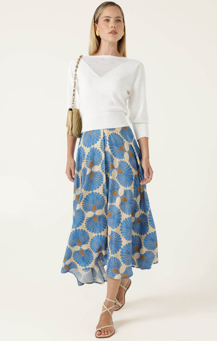 Sacha Drake  modeling a white sleeveless top with a textured chest panel and a high-waisted midi skirt featuring a blue, beige, and brown floral fan pattern, paired with beige strappy heels and holding a small metallic clutch. On a white studio background