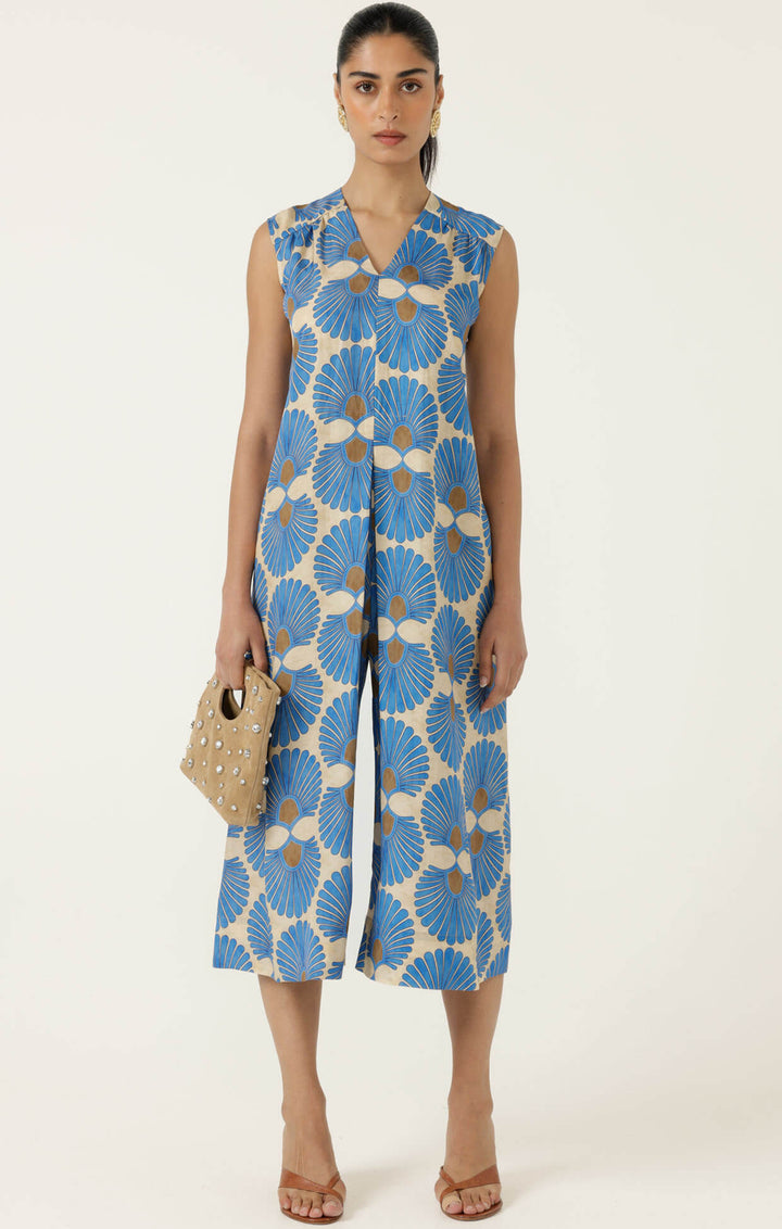 Sacha Drake  models a sleeveless wrap-style jumpsuit with wide cropped legs, featuring a blue and beige geometric floral pattern and a matching waist belt. She is holding a small studded tan handbag and wearing tan heeled sandals against a plain off-white studio background.