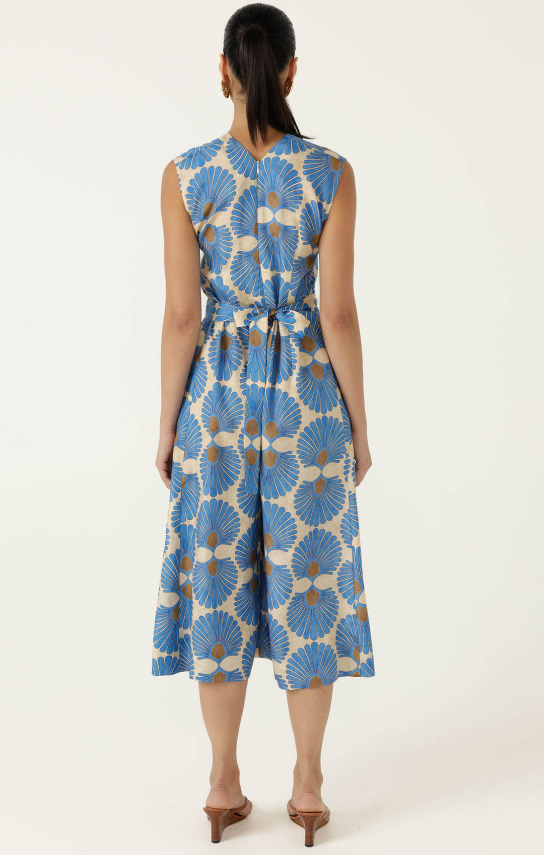 Sacha Drake  models a sleeveless wrap-style jumpsuit with wide cropped legs, featuring a blue and beige geometric floral pattern and a matching waist belt. She is holding a small studded tan handbag and wearing tan heeled sandals against a plain off-white studio background. Back view