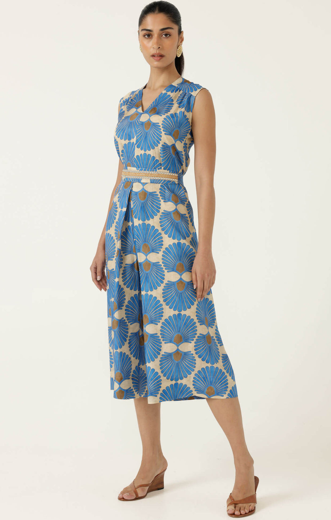 Sacha Drake  models a sleeveless wrap-style jumpsuit with wide cropped legs, featuring a blue and beige geometric floral pattern and a matching waist belt. She is holding a small studded tan handbag and wearing tan heeled sandals against a plain off-white studio background.
