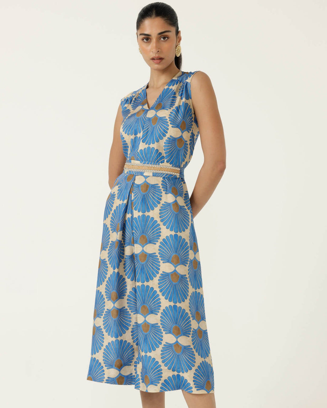 Sacha Drake  models a sleeveless wrap-style jumpsuit with wide cropped legs, featuring a blue and beige geometric floral pattern and a matching waist belt. She is holding a small studded tan handbag and wearing tan heeled sandals against a plain off-white studio background.
