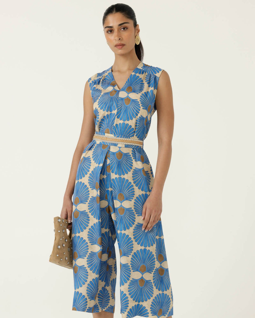 Sacha Drake  models a sleeveless wrap-style jumpsuit with wide cropped legs, featuring a blue and beige geometric floral pattern and a matching waist belt. She is holding a small studded tan handbag and wearing tan heeled sandals against a plain off-white studio background.