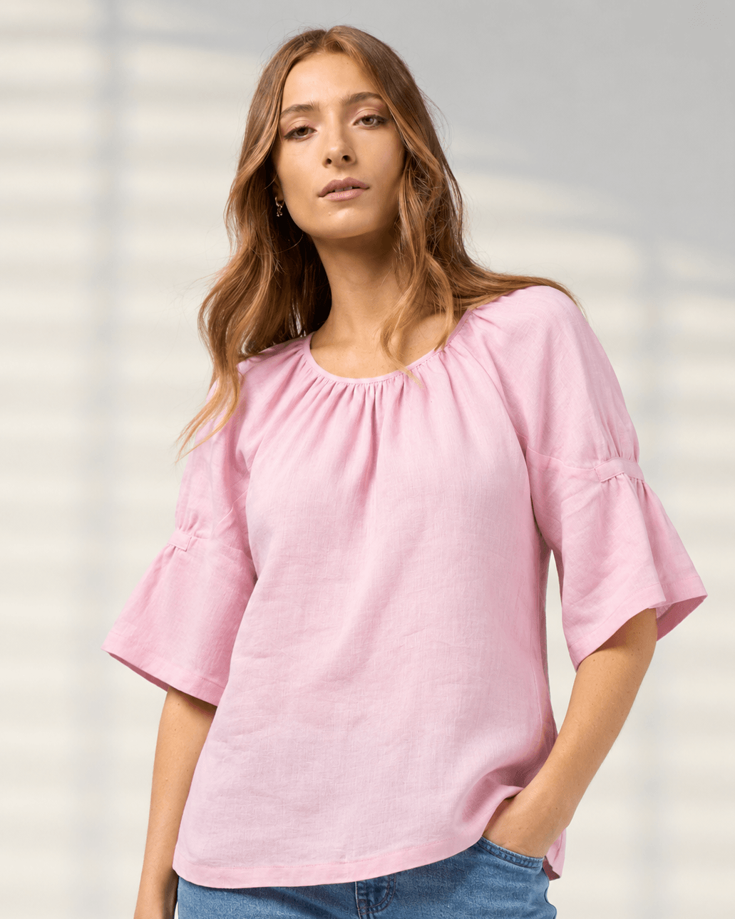 Madly Sweetly model wearing a light pink linen-blend top with a gathered round neckline and flared elbow-length sleeves, paired with blue jeans, set against a soft gray background with subtle leaf shadows.