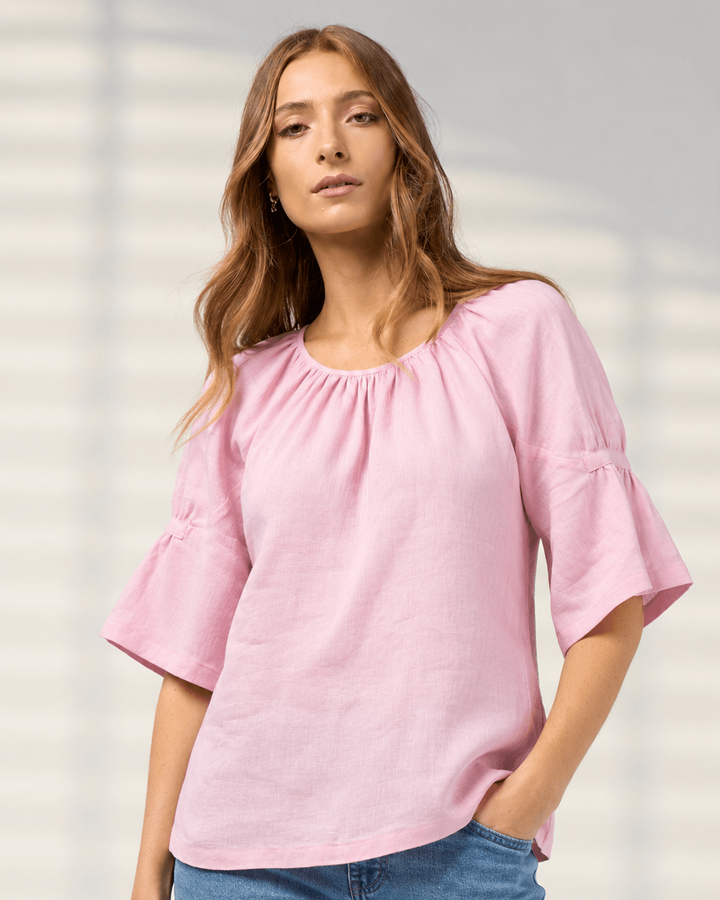 Madly Sweetly model wearing a light pink linen-blend top with a gathered round neckline and flared elbow-length sleeves, paired with blue jeans, set against a soft gray background with subtle leaf shadows.