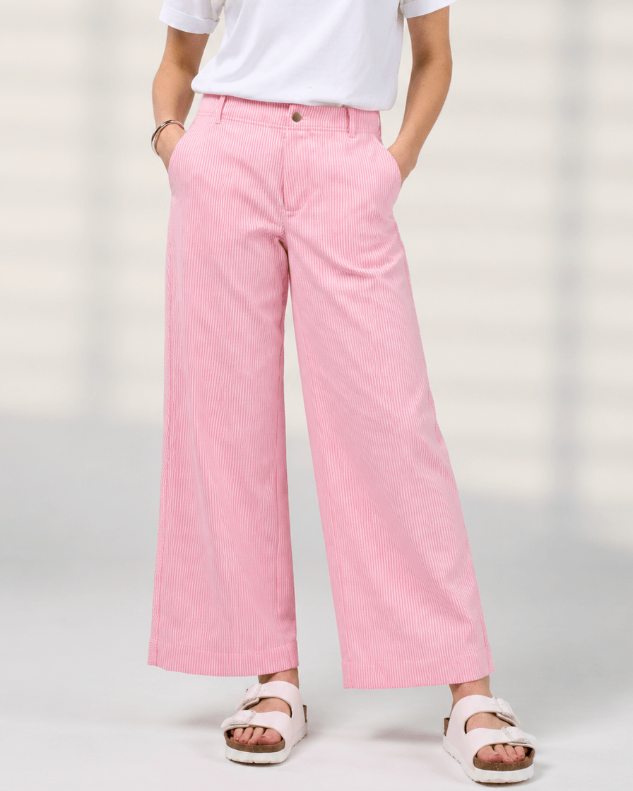 Madly Sweetly model wearing pink wide-leg pinstripe trousers, a white short-sleeve top, and white slide sandals, standing against a light neutral background.