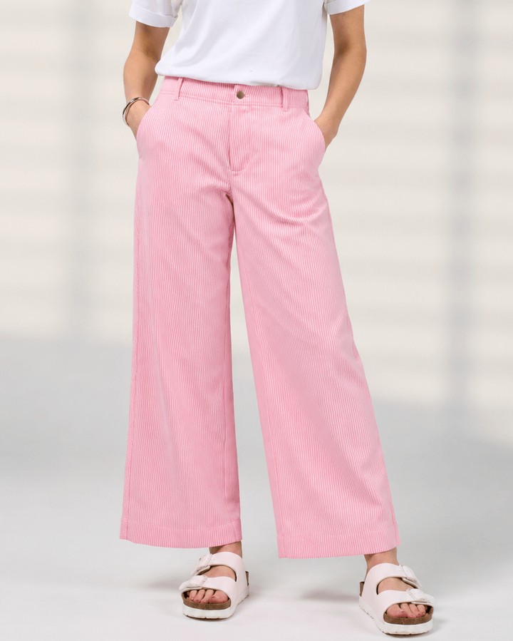 Madly Sweetly model wearing pink wide-leg pinstripe trousers, a white short-sleeve top, and white slide sandals, standing against a light neutral background.