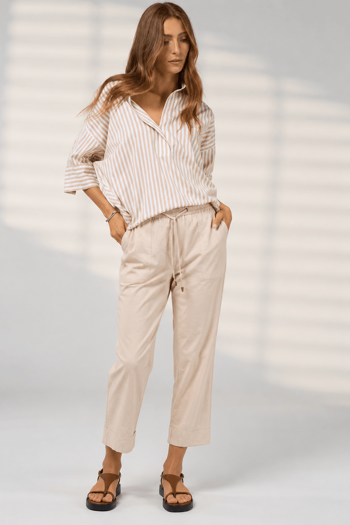 Madly Sweetly model  wearing light beige drawstring cropped pants with a striped shirt and brown sandals, standing against a light gray studio background with soft window-like shadows.