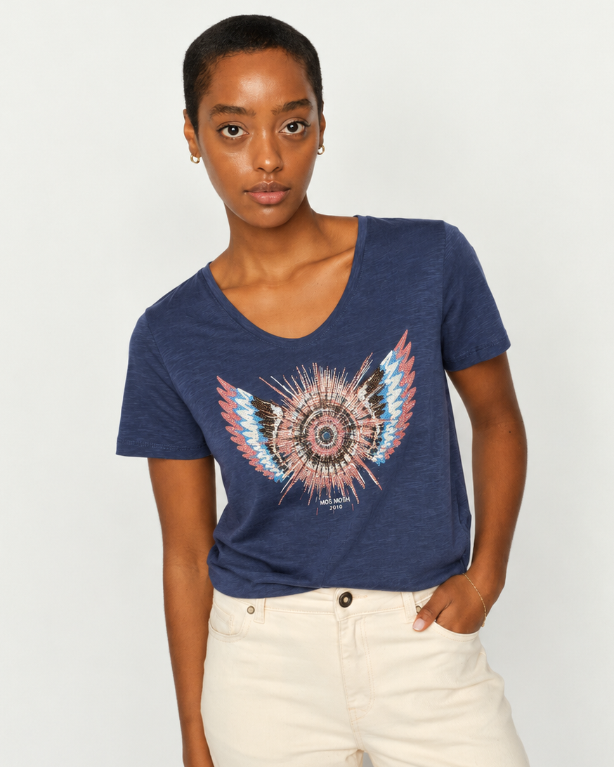 Mos Mosh  woman modeling a navy blue short-sleeve t-shirt with a colorful feathered wing graphic on the front, paired with cream-colored pants, against a plain light gray background.
