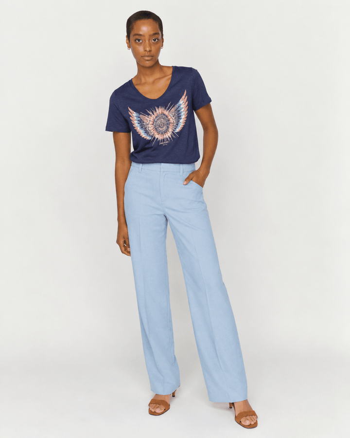 Mos Mosh model stands against a light gray studio background wearing a navy blue t-shirt with a pink and white wing graphic, paired with light blue wide-leg trousers and brown sandals.