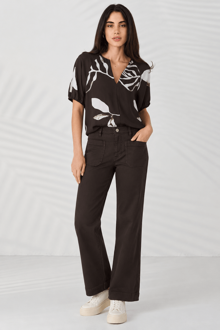 Lania The Label Model modeling a short-sleeve button-front blouse with a dark brown base and large white botanical leaf print, paired with matching dark pants, against a light gray studio backdrop with soft leaf shadows.