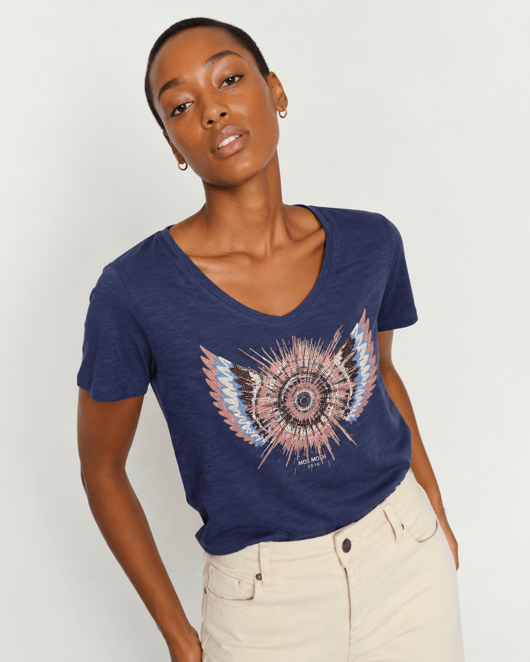 Mos Mosh model wearing a navy blue V-neck t-shirt with a colorful feathered mandala graphic on the front, paired with light beige pants, against a plain light gray background.