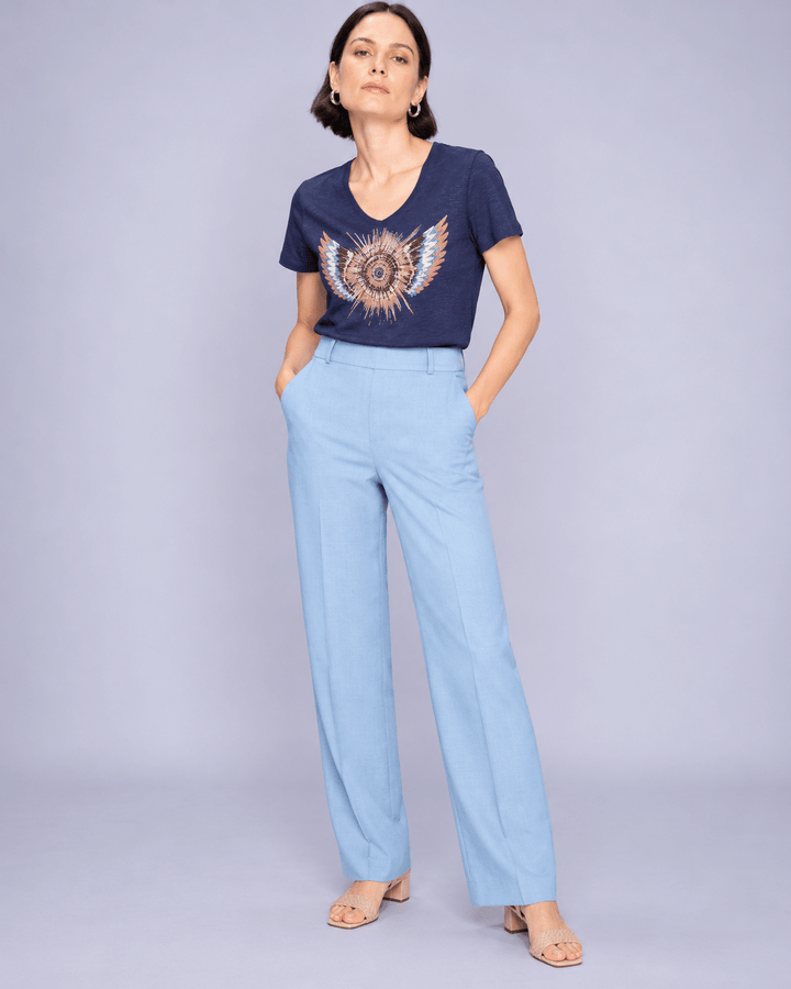 Mos Mosh  woman modeling a navy blue t-shirt with a gold wing graphic and light blue wide-leg trousers, standing against a solid light gray studio background.