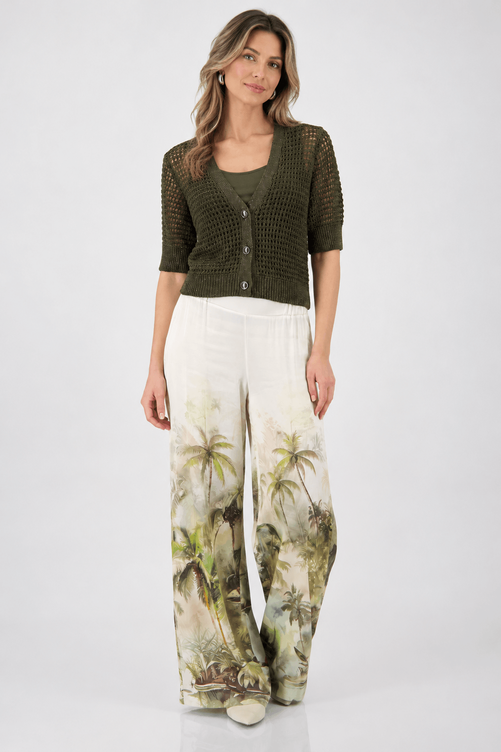 Monari woman modeling a short-sleeve olive green knit cardigan over a matching olive green top, paired with wide-leg white pants featuring a green and beige palm tree print, against a light gray studio background.