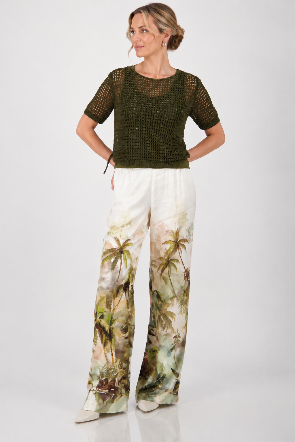 Monari  modeling a short-sleeve olive green knit top paired with white wide-leg pants featuring a green palm tree print, set against a plain light gray studio background.