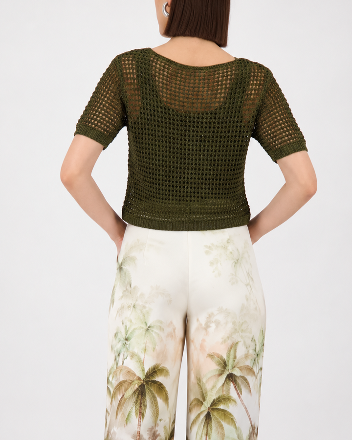 Monari  modeling a short-sleeve olive green knit top paired with white wide-leg pants featuring a green palm tree print, set against a plain light gray studio background.
