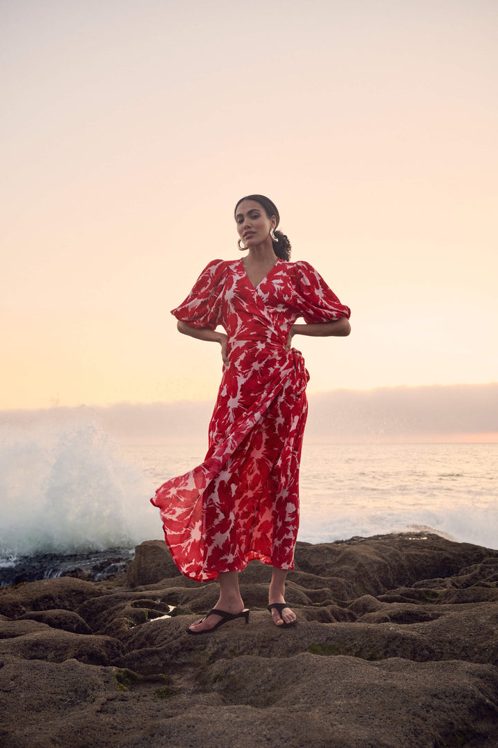Pom Amsterdam model wearing a red and white abstract floral print wrap dress, standing on a rocky beach with the ocean and sunset in the background.