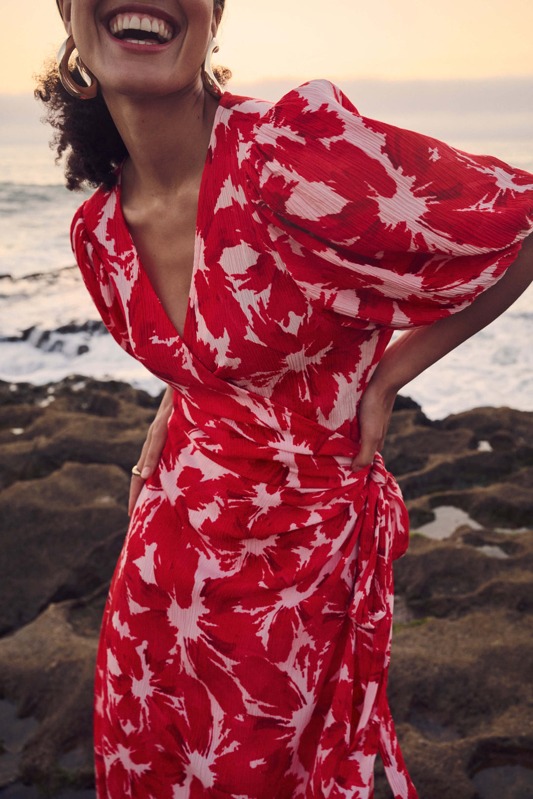 Pom Amsterdam model wearing a red and white abstract floral print wrap dress, standing on a rocky beach with the ocean and sunset in the background.