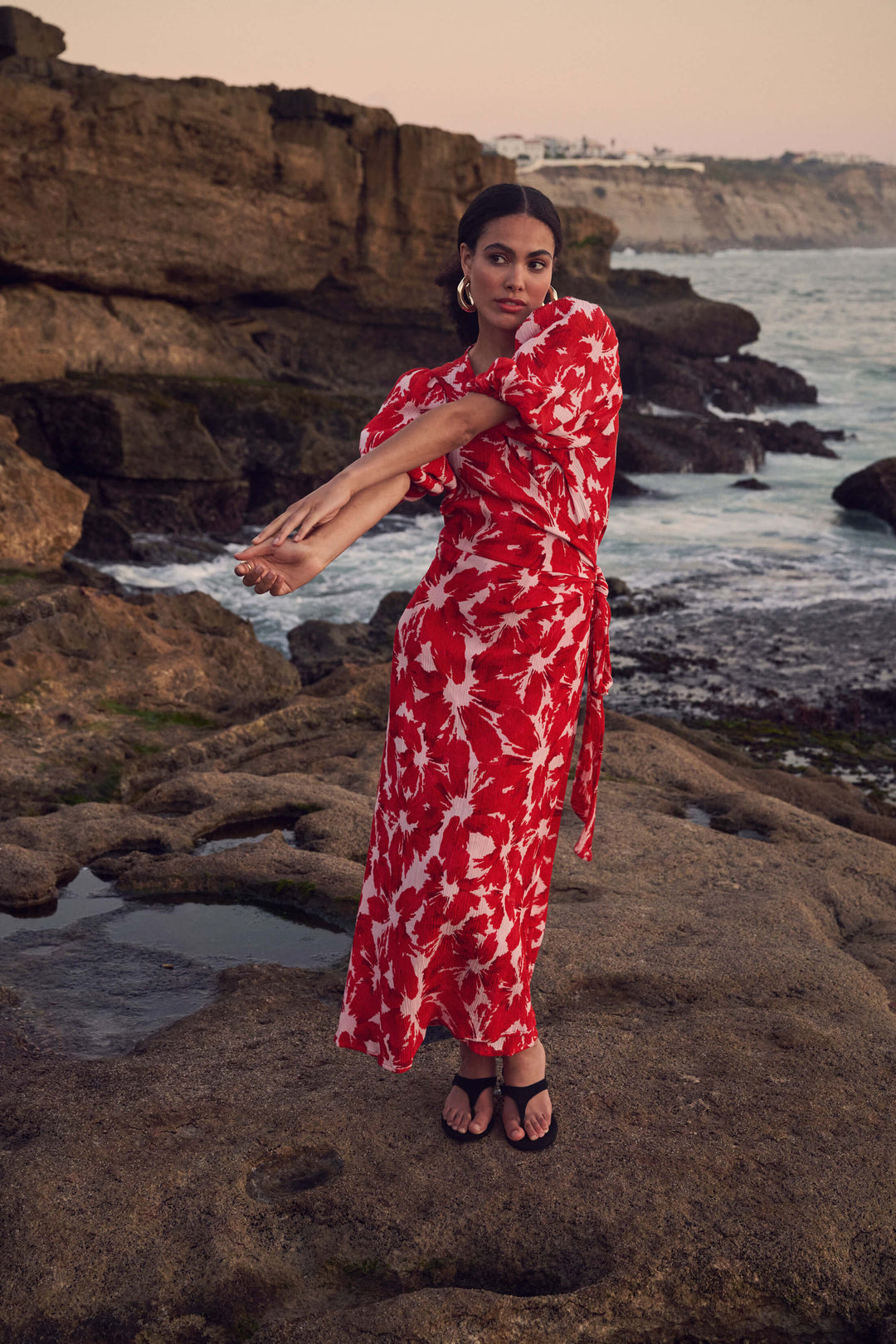 Pom Amsterdam model wearing a red and white abstract floral print wrap dress, standing on a rocky beach with the ocean and sunset in the background.