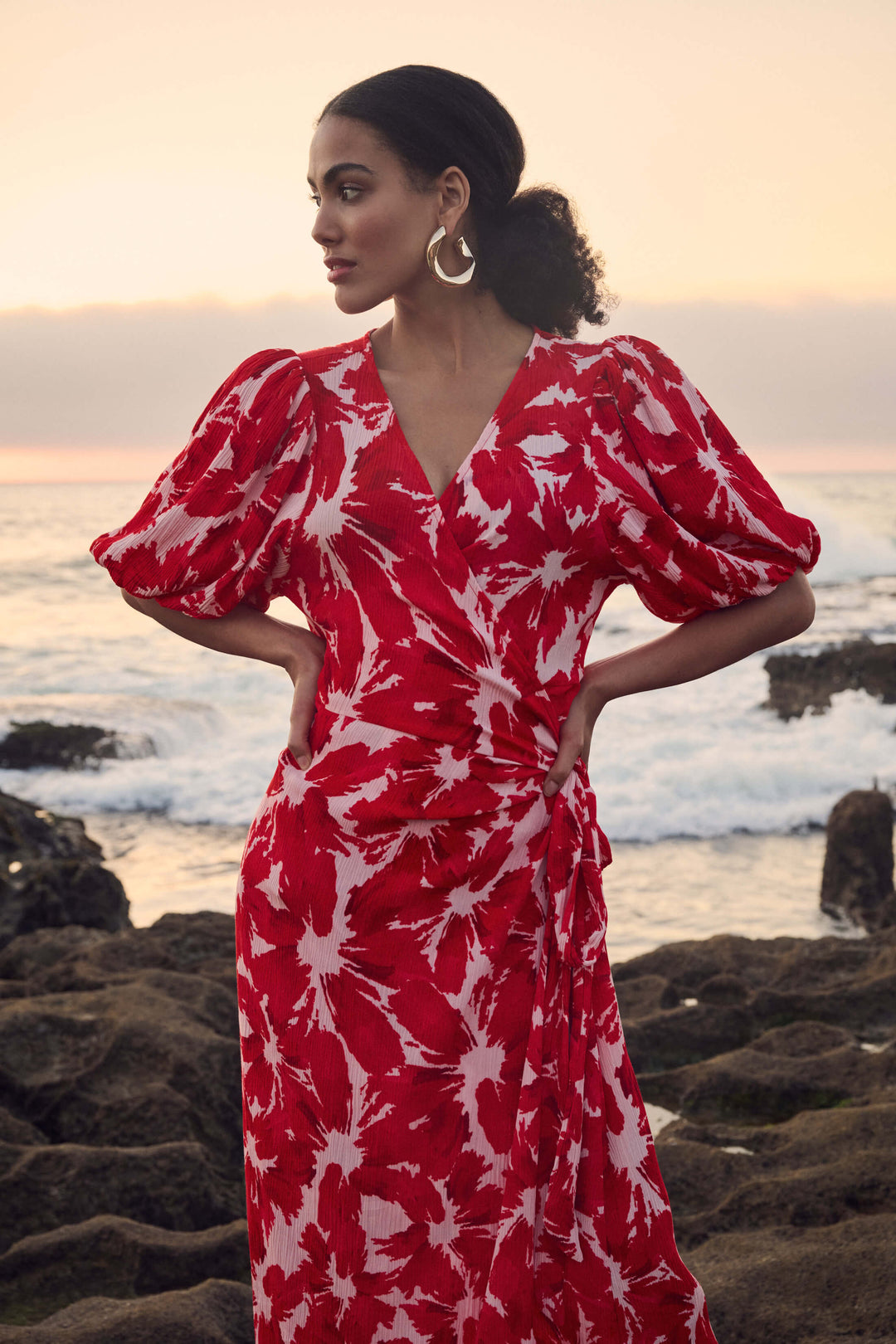 Pom Amsterdam model wearing a red and white abstract floral print wrap dress, standing on a rocky beach with the ocean and sunset in the background.