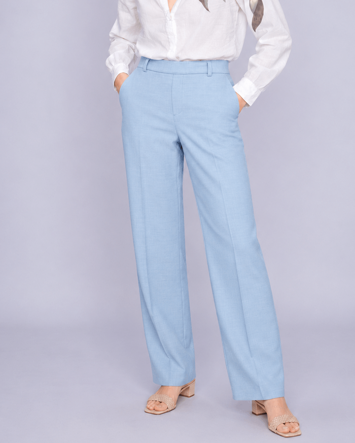 Mos Mosh woman modeling a white long-sleeve blouse with metallic gold leaf embroidery on the shoulders, paired with light blue wide-leg trousers and beige sandals, against a solid light gray studio background.