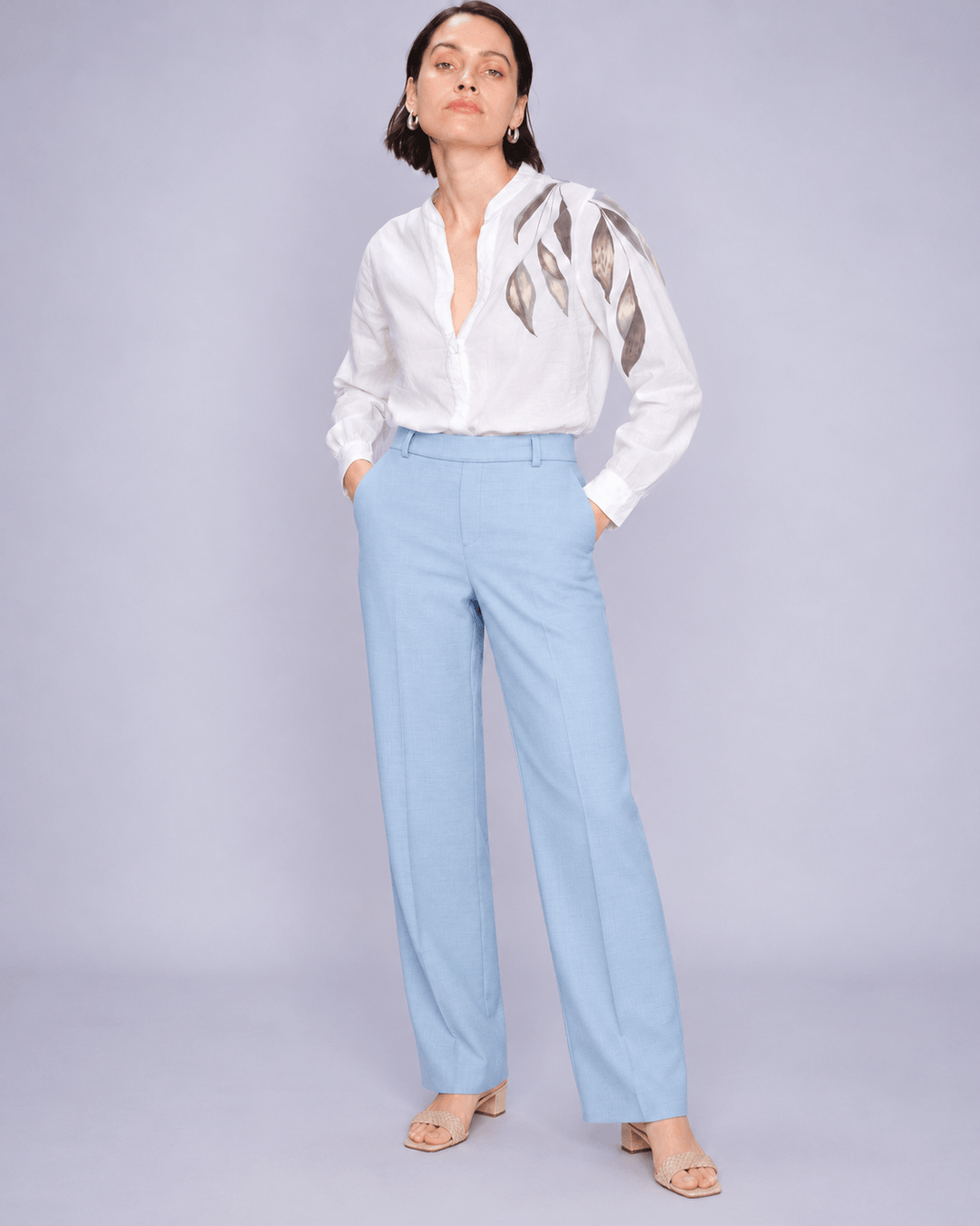 Mos Mosh woman modeling a white long-sleeve blouse with metallic gold leaf embroidery on the shoulders, paired with light blue wide-leg trousers and beige sandals, against a solid light gray studio background.