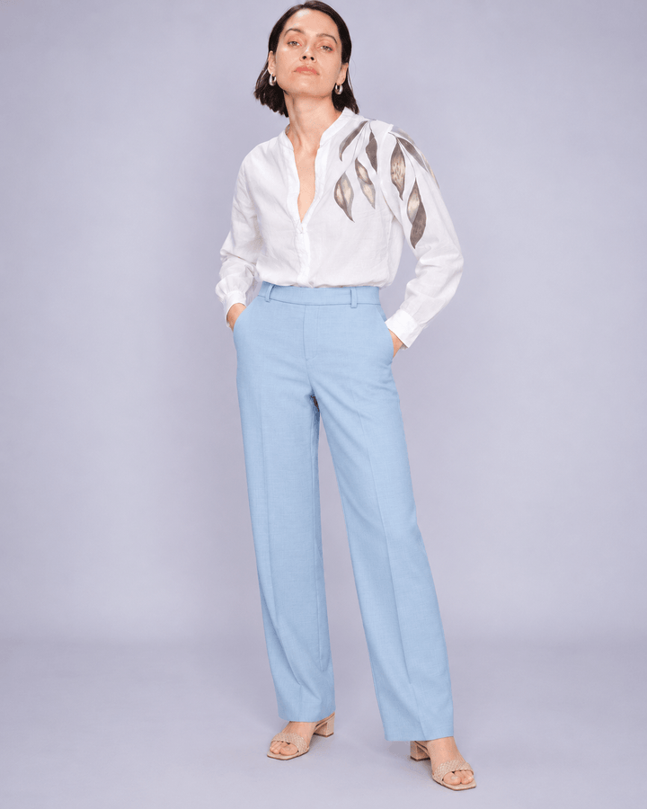 Mos Mosh woman modeling a white long-sleeve blouse with metallic gold leaf embroidery on the shoulders, paired with light blue wide-leg trousers and beige sandals, against a solid light gray studio background.