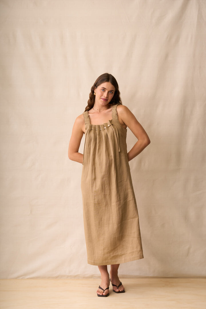 Neris modeling a sleeveless, midi-length linen dress in a warm taupe-brown color, standing against a neutral beige fabric backdrop.