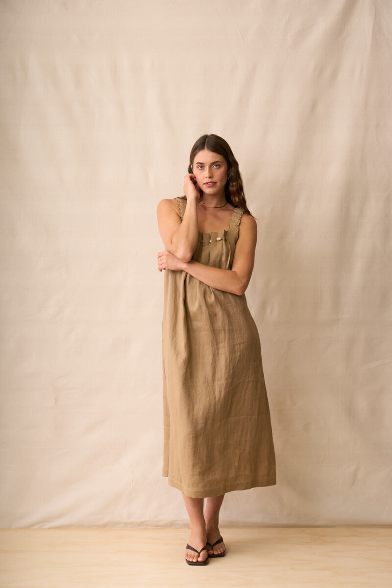 Neris modeling a sleeveless, midi-length linen dress in a warm taupe-brown color, standing against a neutral beige fabric backdrop.