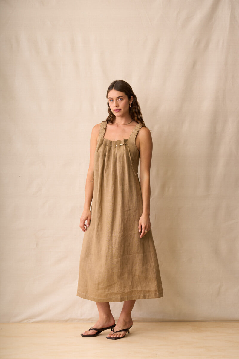 Neris modeling a sleeveless, midi-length linen dress in a warm taupe-brown color, standing against a neutral beige fabric backdrop.