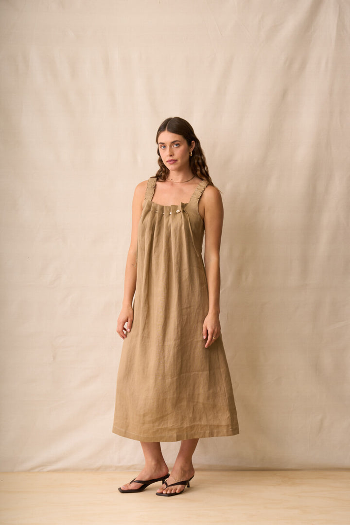 Neris modeling a sleeveless, midi-length linen dress in a warm taupe-brown color, standing against a neutral beige fabric backdrop.