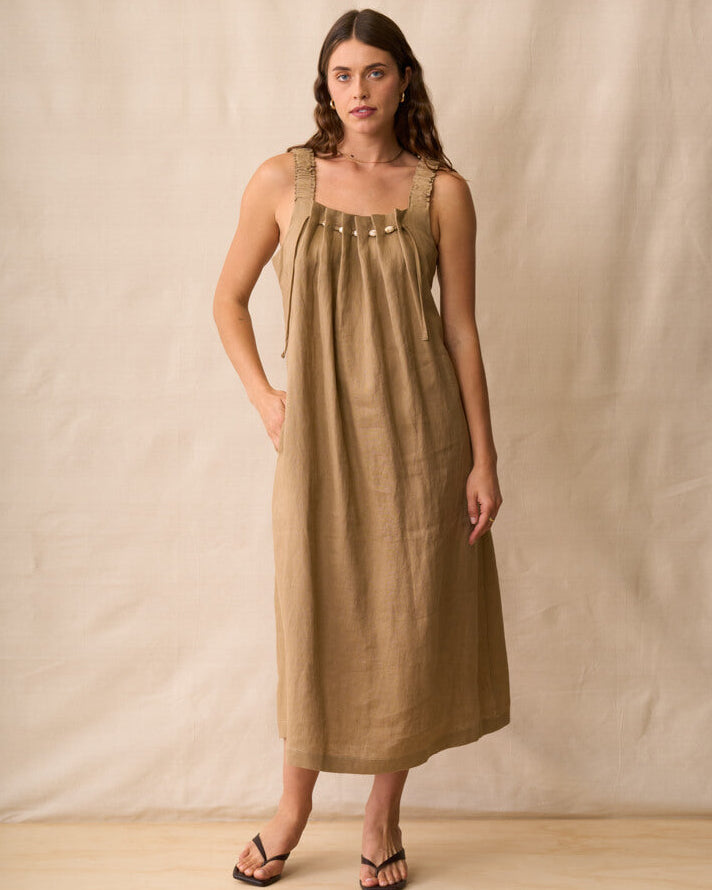 Neris modeling a sleeveless, midi-length linen dress in a warm taupe-brown color, standing against a neutral beige fabric backdrop.