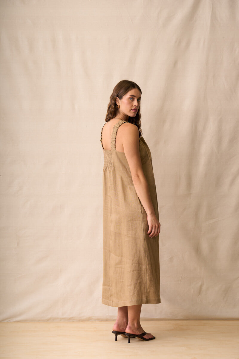 Neris modeling a sleeveless, midi-length linen dress in a warm taupe-brown color, standing against a neutral beige fabric backdrop.