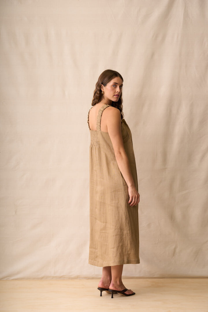Neris modeling a sleeveless, midi-length linen dress in a warm taupe-brown color, standing against a neutral beige fabric backdrop.