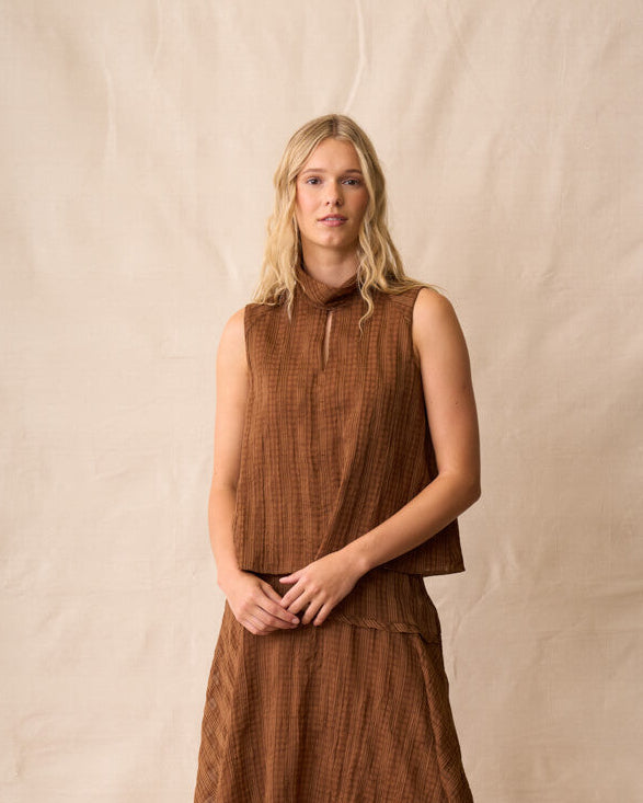 Neris model  stands against a beige fabric backdrop wearing a sleeveless brown textured top, dark blue denim shorts, and brown sandals.