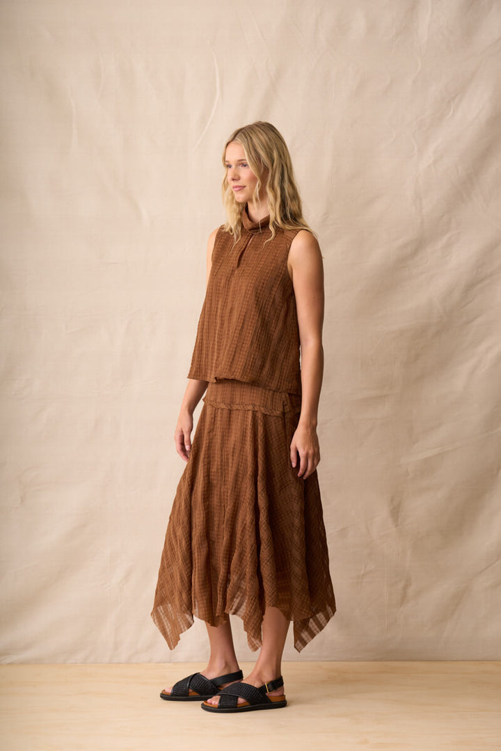 Neris model  stands against a beige fabric backdrop wearing a sleeveless brown textured top, dark blue denim shorts, and brown sandals.