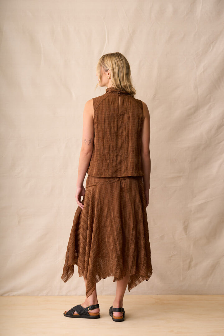 Neris model  stands against a beige fabric backdrop wearing a sleeveless brown textured top, dark blue denim shorts, and brown sandals.