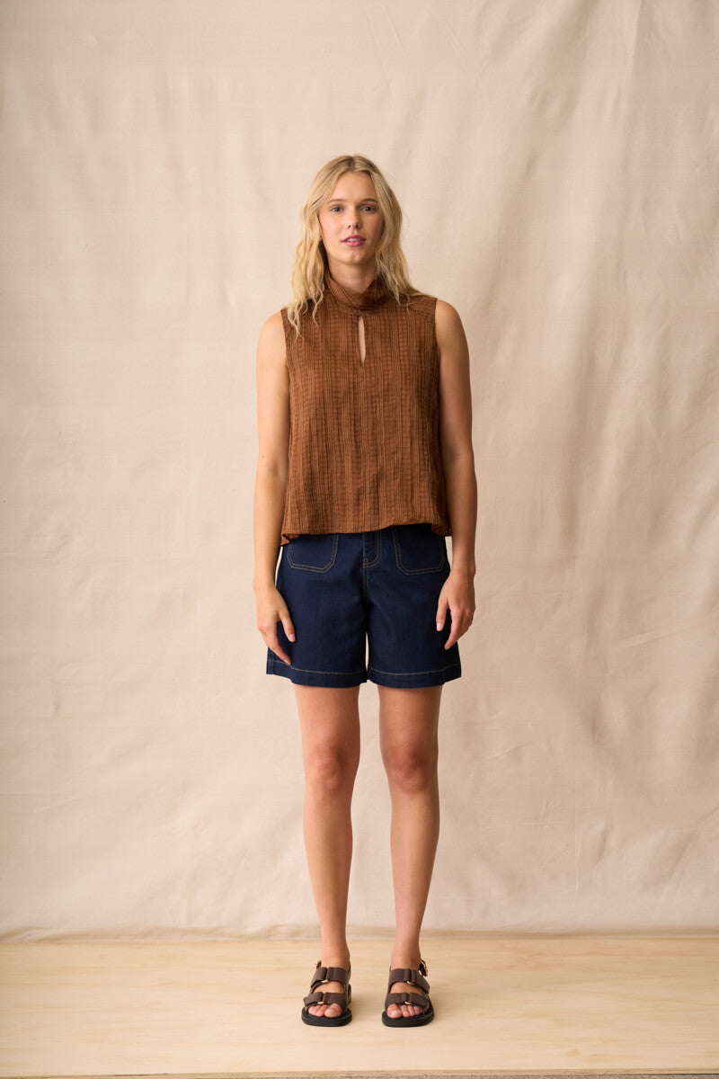Neris model  stands against a beige fabric backdrop wearing a sleeveless brown textured top, dark blue denim shorts, and brown sandals.