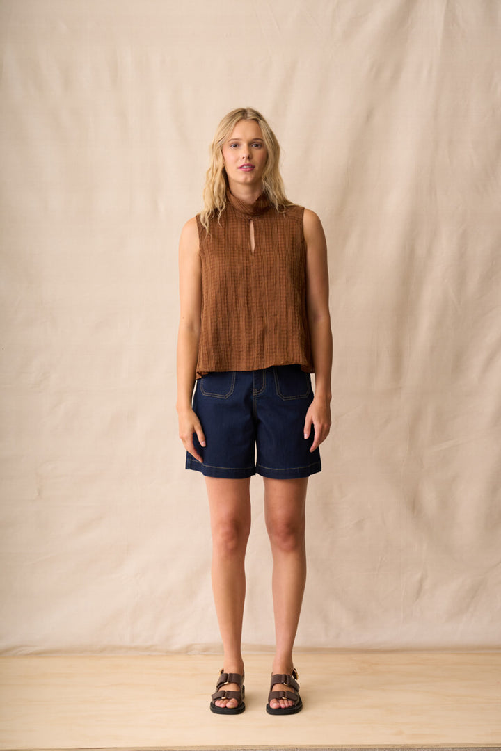 Neris model  stands against a beige fabric backdrop wearing a sleeveless brown textured top, dark blue denim shorts, and brown sandals.