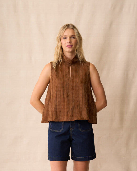 Neris model  stands against a beige fabric backdrop wearing a sleeveless brown textured top, dark blue denim shorts, and brown sandals.