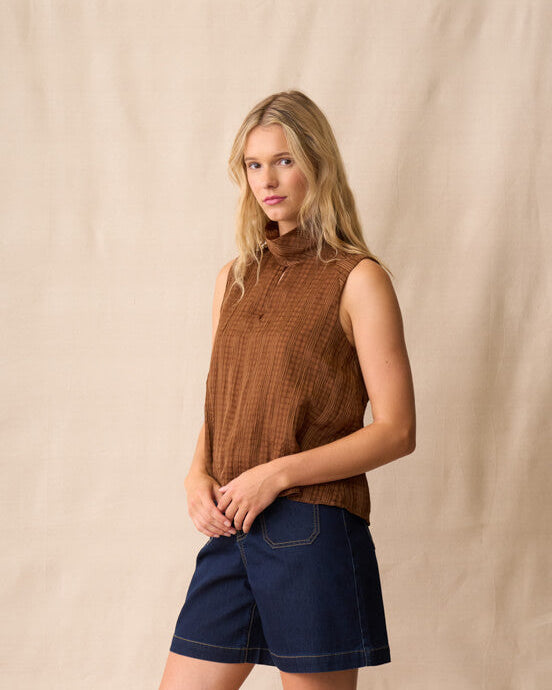 Neris model  stands against a beige fabric backdrop wearing a sleeveless brown textured top, dark blue denim shorts, and brown sandals.