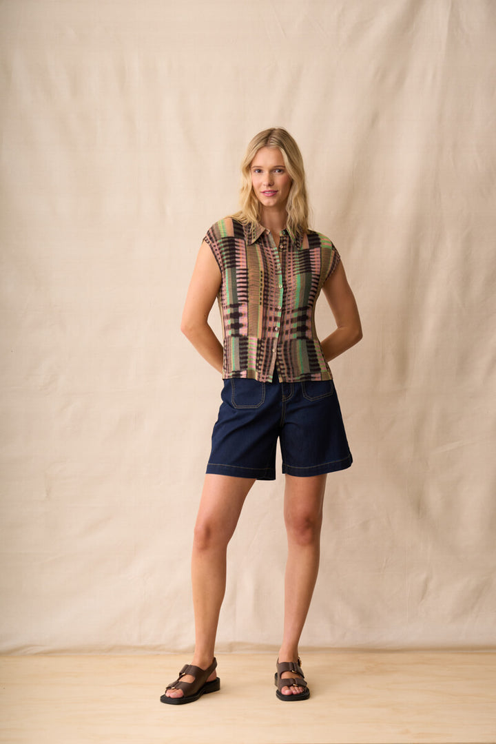 Neris model stands against a beige fabric backdrop wearing a short-sleeve button-up shirt with a multicolor geometric stripe pattern, navy blue shorts, and dark sandals.