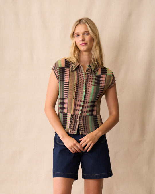 Neris model stands against a beige fabric backdrop wearing a short-sleeve button-up shirt with a multicolor geometric stripe pattern, navy blue shorts, and dark sandals.