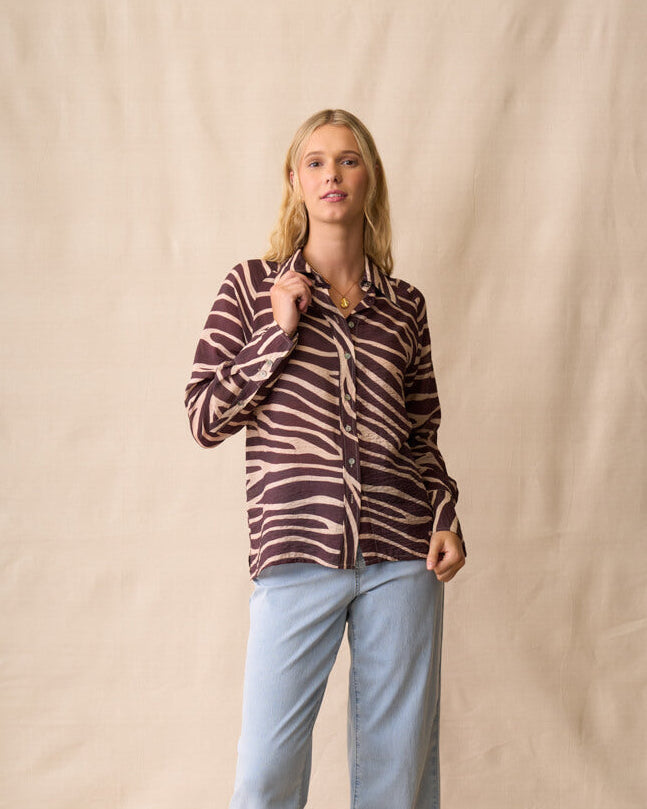 Neris modeling a long-sleeve button-up shirt with a dark brown and beige zebra-like stripe pattern, paired with light blue jeans and dark sandals, standing against a beige fabric backdrop.