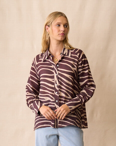Neris modeling a long-sleeve button-up shirt with a dark brown and beige zebra-like stripe pattern, paired with light blue jeans and dark sandals, standing against a beige fabric backdrop.
