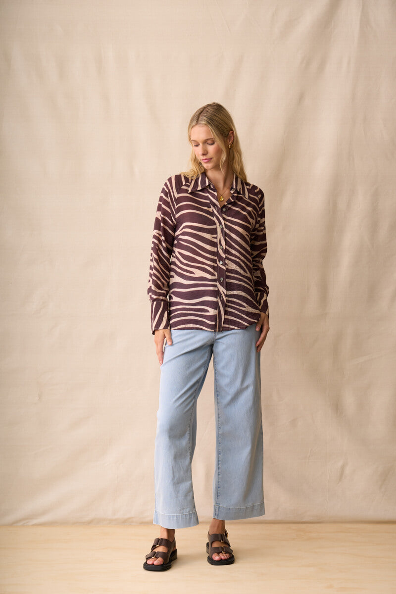 Neris modeling a long-sleeve button-up shirt with a dark brown and beige zebra-like stripe pattern, paired with light blue jeans and dark sandals, standing against a beige fabric backdrop.