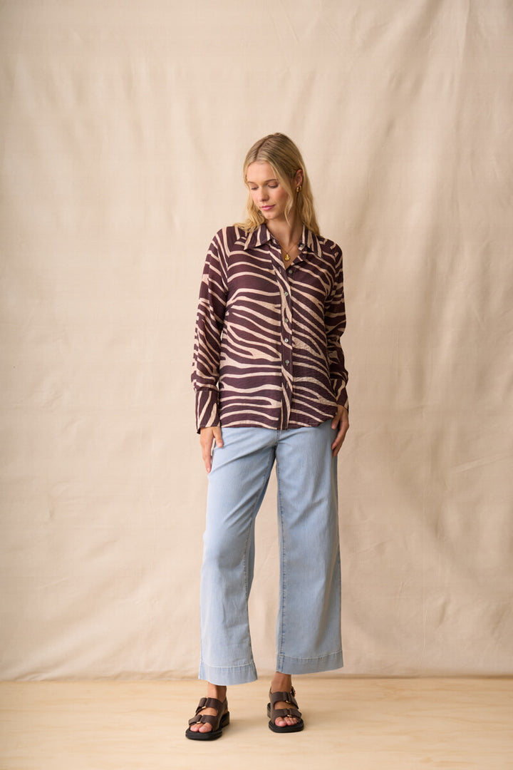 Neris modeling a long-sleeve button-up shirt with a dark brown and beige zebra-like stripe pattern, paired with light blue jeans and dark sandals, standing against a beige fabric backdrop.