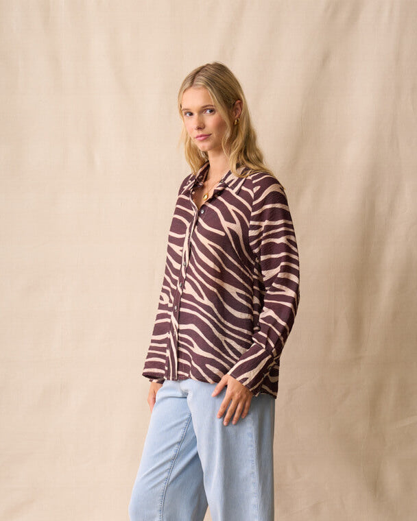 Neris modeling a long-sleeve button-up shirt with a dark brown and beige zebra-like stripe pattern, paired with light blue jeans and dark sandals, standing against a beige fabric backdrop.