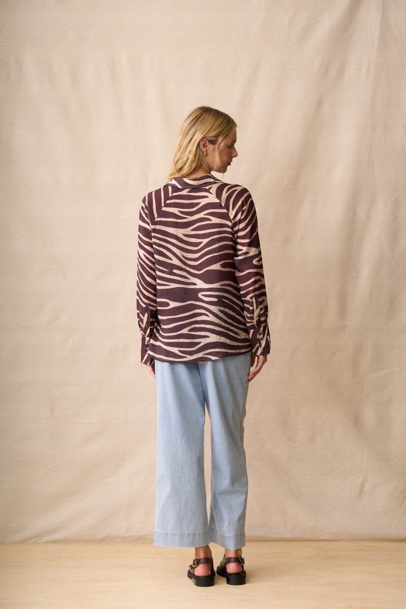 Neris modeling a long-sleeve button-up shirt with a dark brown and beige zebra-like stripe pattern, paired with light blue jeans and dark sandals, standing against a beige fabric backdrop. Back View.